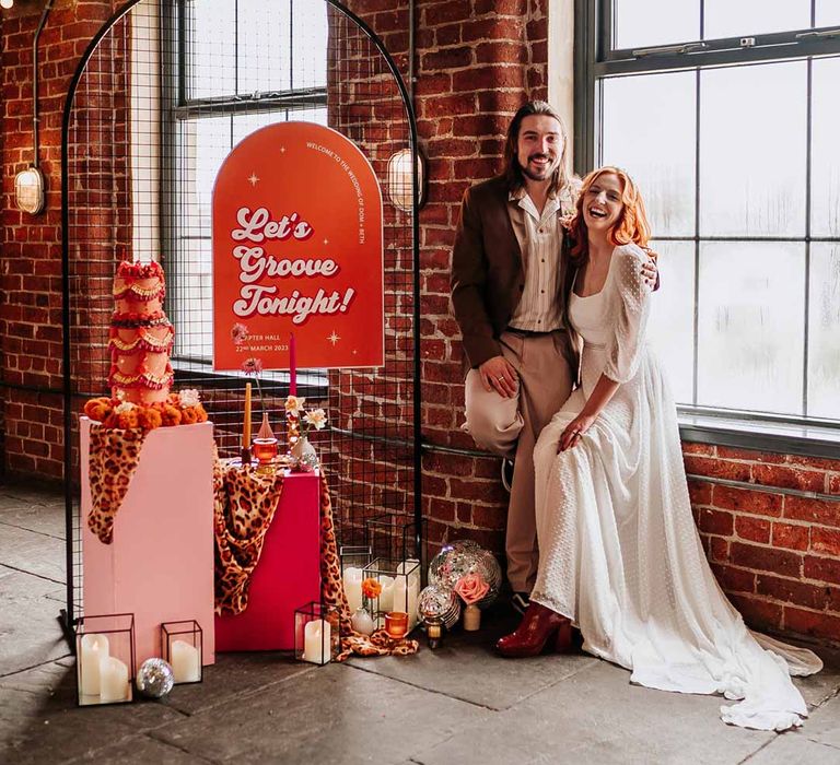 Bride in three quarter length puff sheer sleeve wedding dress with delicate polka dot overlay and square neck posing with groom in brown blazer, white shirt, grey suit trousers and vans shoes next to retro wedding decor