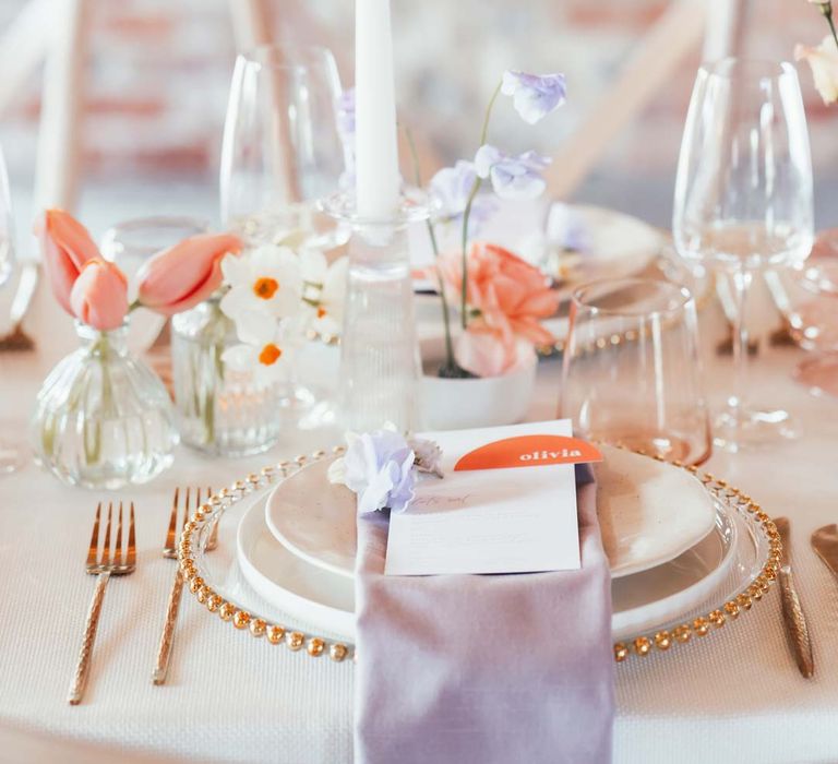 White tapered candle in clear candlestick holder on peach and lilac wedding tablescape with lilac wedding napkin and gold crockery 