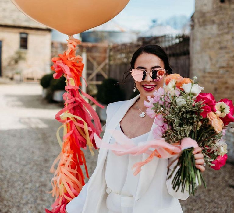 Bride in white bridal suit with rose gold wedding sunglasses holding large mixed seasonal flower bridal bouquet and large balloon with wedding streamers attached