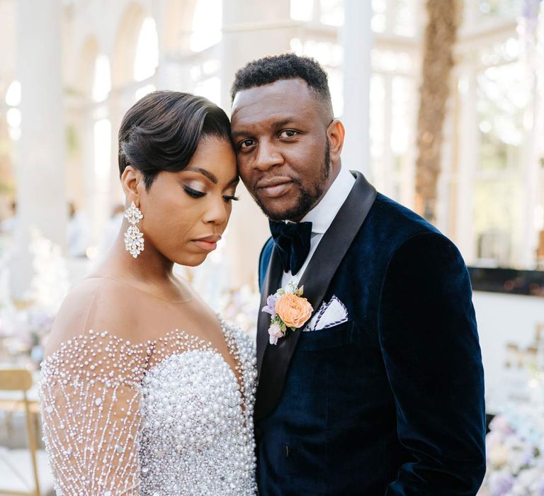 Bride in illusion v-neck sheer off shoulder long sleeve pearl embellished wedding gown standing with groom in velvet midnight blue double breasted groom’s blazer, velvet bowtie and peach garden rose boutonniere at vow renewal at Syon Park wedding venue