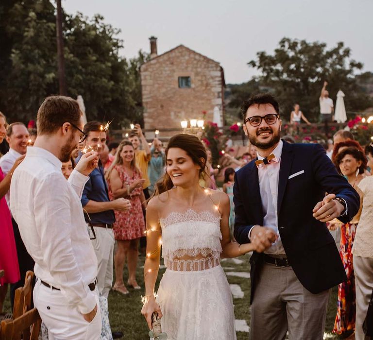 groom in grey chinos navy blazer and orange bow tie holding hands with his bride in a strapless lace wedding dress and fairy lights at greece destination wedding