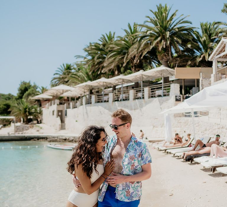 Bride in white swimsuit strolls along beach with groom in floral shirt and blue swim shorts at destination wedding in Hvar Croatia