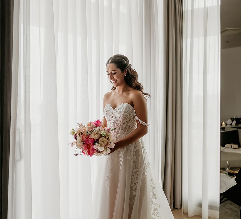 Brride in 3D lace applique off-the-shoulder wedding dress with curled half up half down hair holds a pink and blush wedding bouquet