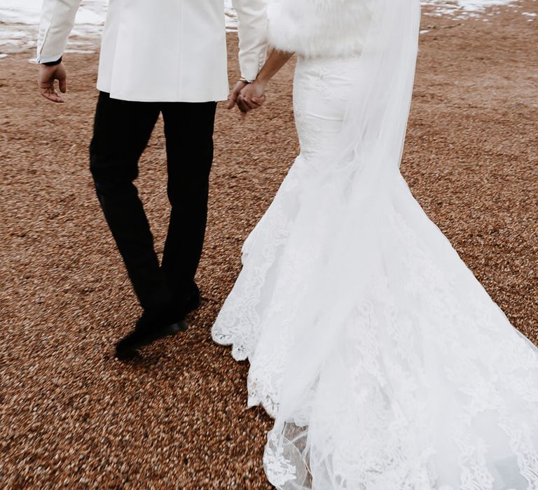 Bride in fishtail wedding gown with faux fur stole for winter wedding walking with groom in white tuxedo 