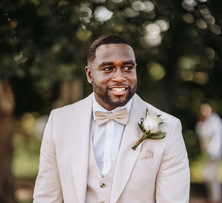 Groom wearing cream three piece wedding suit with champagne bow tie and pocket square with white rose buttonhole 