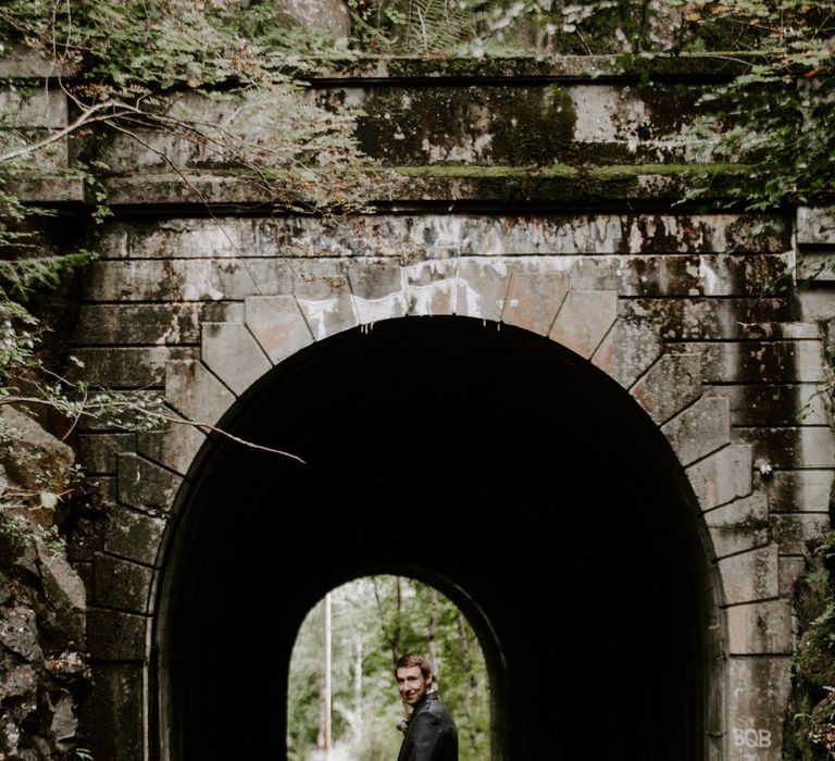 Groom in traditional Scottish kilt and dress stands under a bridge