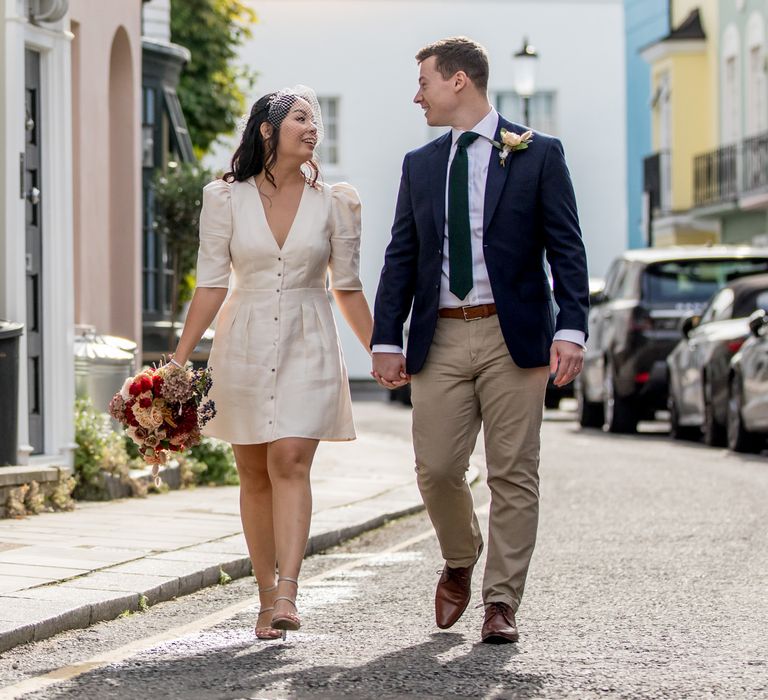 Bride in short wedding dress with button front holding hands with her groom in a navy blazer 