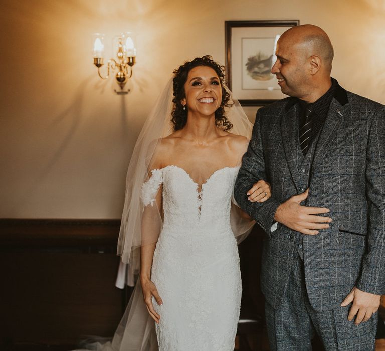 Bride with brown curly hair in a strapless Pronovias wedding dress standing arm in arm with her father in a grey wool suit 