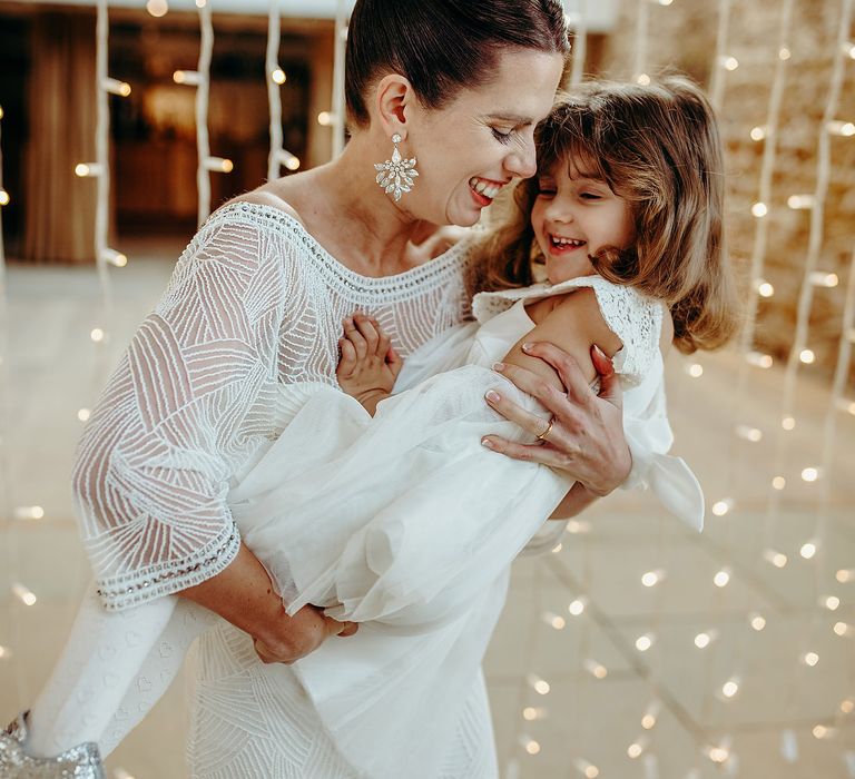 Bride holds flower girl with fairy lights curtain twinkling behind 