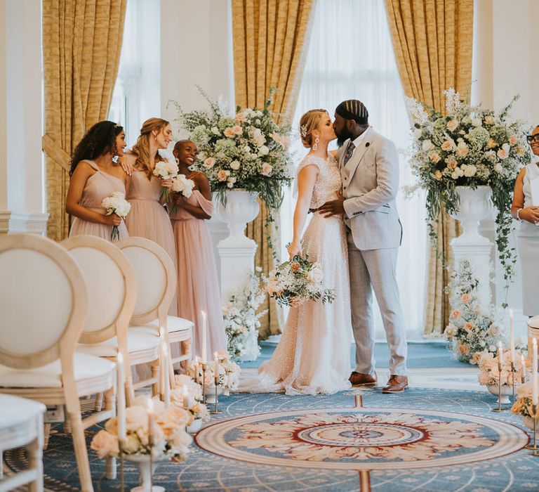 Bride in a sparkly dress and the groom in a grey suit kissing at the altar with the wedding celebrant and bridesmaids standing close by 