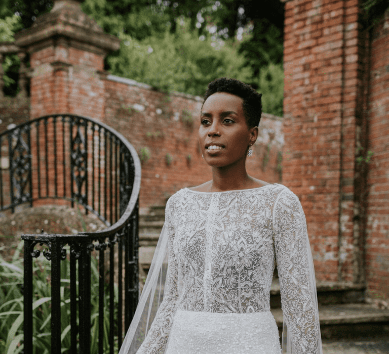 Black bride with cropped hair wears a high neck long sleeve wedding dress. She walking down steps and the cape of the dress flows behind her.