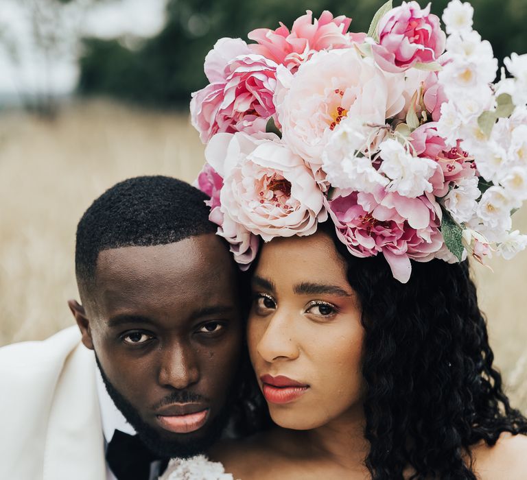 A Black couple pose for wedding photos. He wears a white tuxedo and she a long sleeved white dress. She has a large pink flower crown in her loose curly hair.