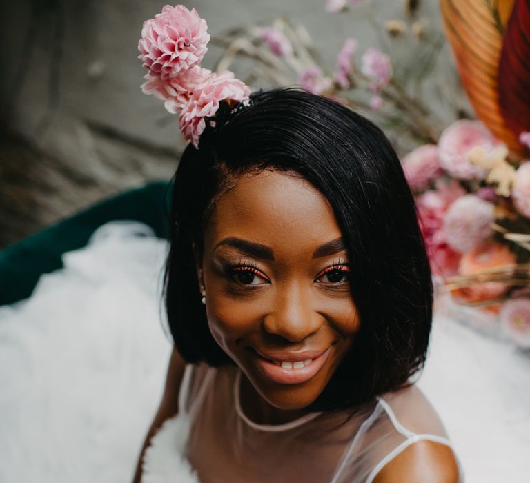 A Black bride with short relaxed hair looks up at the camera smiling. She wears a white dress and holds pink flowers.