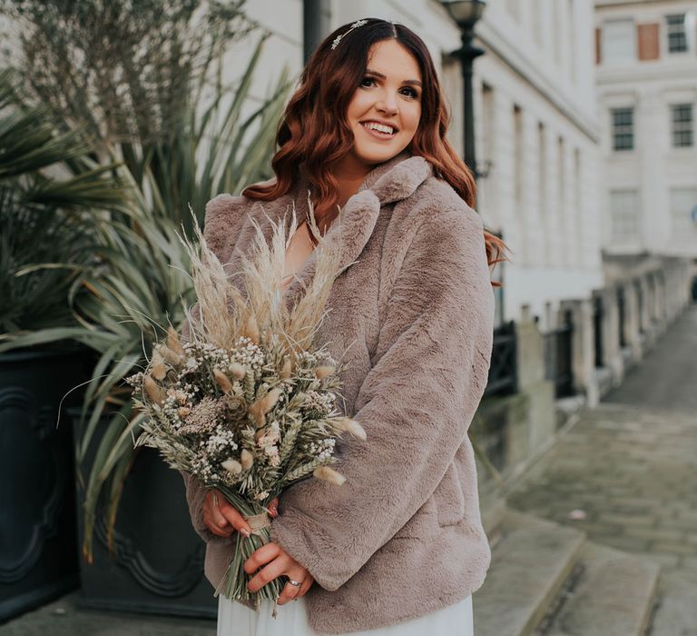 Smiling bride in fur coat holds dried flower bouquet 