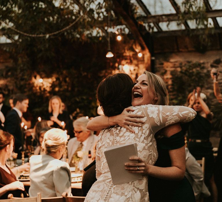 Bride in a long sleeve lace wedding dress by Hermione de Paula hugging her bridesmaid at the wedding reception 