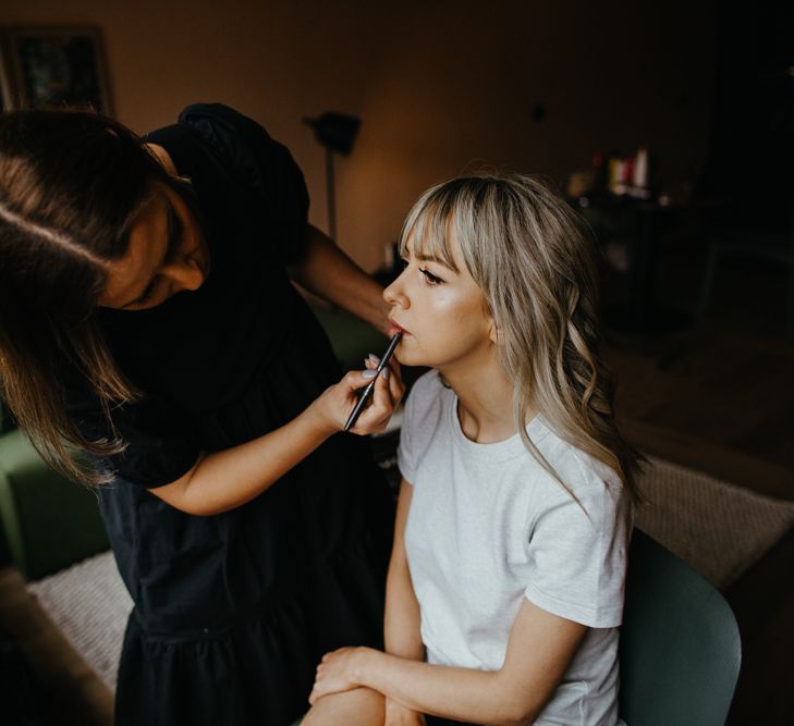 White bride with fringe and wavy blonde hair having lip liner applied by makeup artist