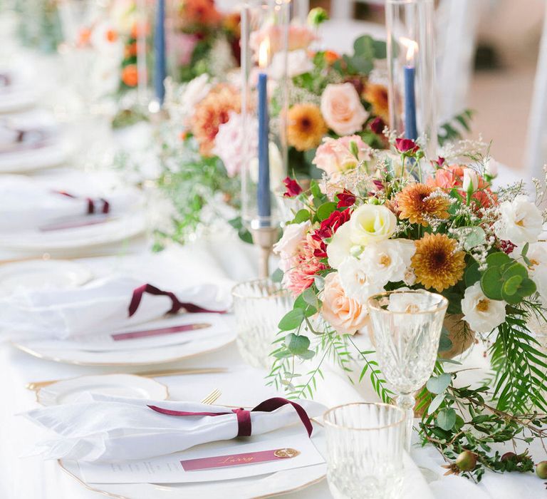 White table place settings with burgundy details, autumnal floral centrepieces and blue tapered candles