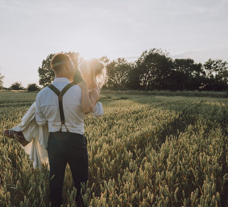 Groom carries bride through field as the sunsets for post-wedding photoshoot 