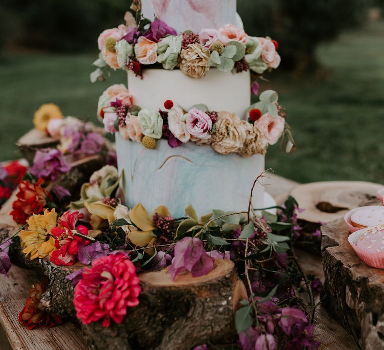 White, blue and pink three tiered wedding cake with multicoloured rose and foliage decor on wooden bark-lined stand with pink and white cupcakes at enchanted forest wedding in Italy