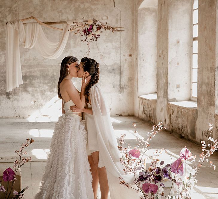 Two brides kissing at romantic ceremony at industrial wedding venue with purple flowers and drift wood hanging installation