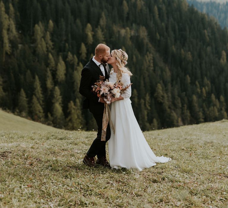 Boho bride at intimate elopement holding an autumn bouquet kissing her groom on top of the Dolomites mountains 