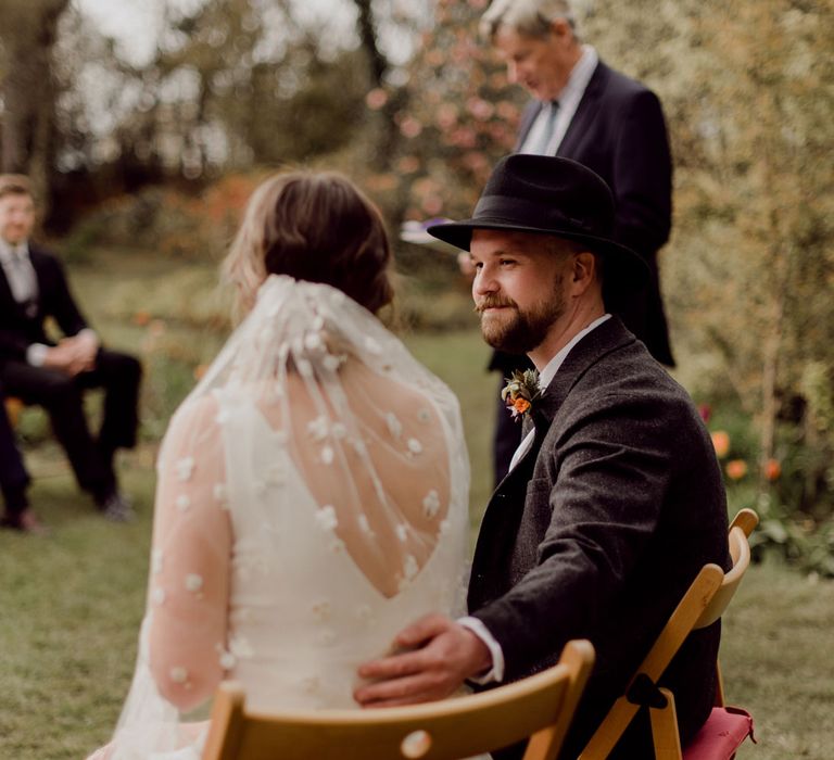 Groom sat wearing grey tweed suit and black fedora smiles with arm resting on the back of bride sat wearing Charlie Brear wedding dress and daisy applique veil