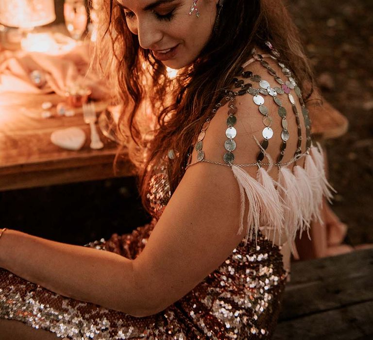 Bride with long curled hair and face glitter smiles wearing rose gold sequinned wedding dress and silver disc shoulder covering sat outside at festival themed wedding