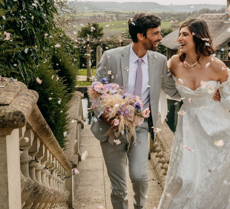 Bride & groom laugh together and walk as groom holds floral bouquet