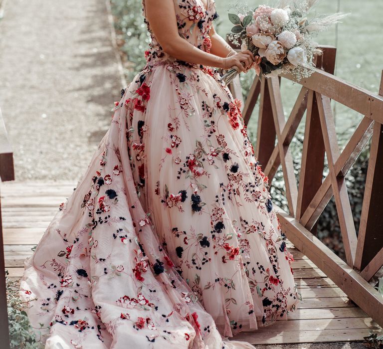 Bride stands on wooden bridge whilst wearing floral appliqué wedding down carrying floral bouquet