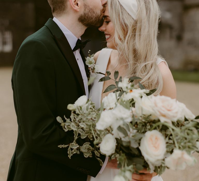 Groom kisses blonde bride on her forehead as she holds white floral bouquet