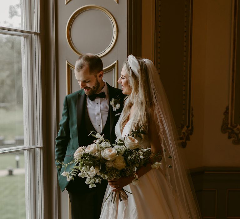 Bride & groom stand beside window on their wedding day as they smile and hold their arms around one another