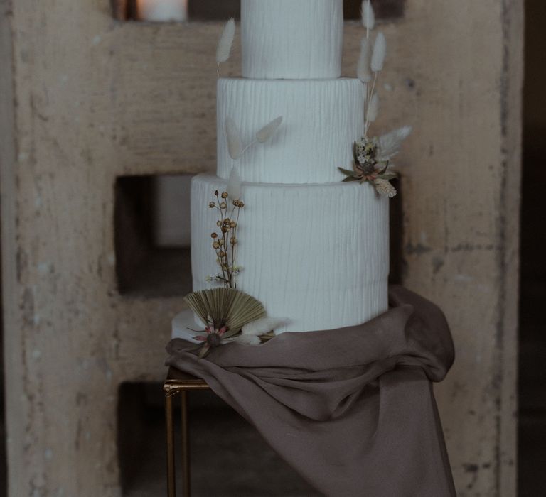 A white, textured three tier wedding cake is decorated with dry grasses and bunny ears for a simple elegant wedding.