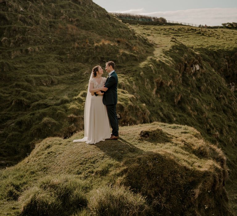 Bride in lace top wedding dress with satin skirt and veil embraces groom on clifftop after Dunluce Castle wedding