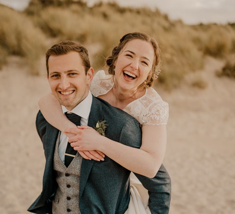 Smiling bride in lace top capped sleeve wedding dress has a piggyback on groom in navy suit on the beach after Dunluce Castle wedding