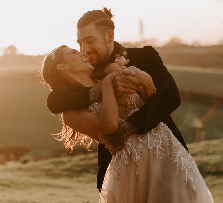 Groom in a black suit embracing his bride in a lace and tulle Berta bridal gown during sunset at Botley Hill Barn wedding venue 