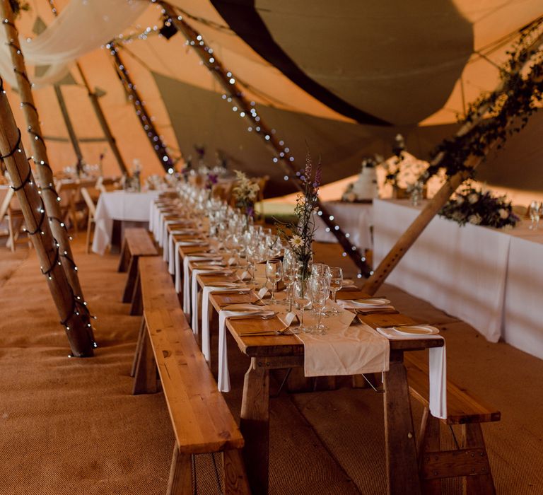 Long wooden rustic wedding breakfast table with white table runner inside tipi with fairy lights, drapery and hessian floor