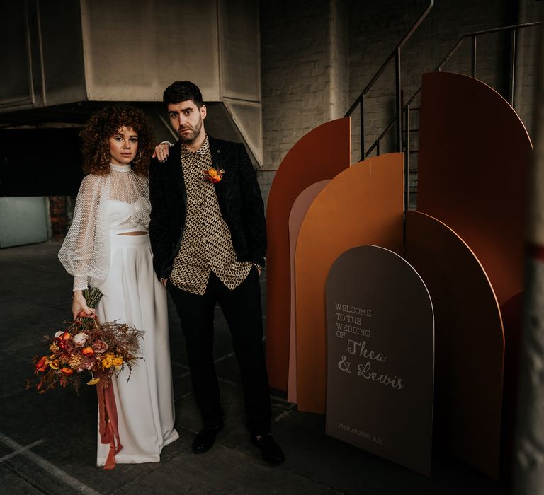 Bride in a jumpsuit holding an autumnal bouquet standing next to her groom in a patterned shirt and geometric boards and wedding sign