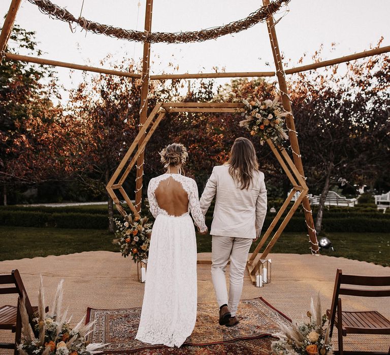 Bride in a keyhole back lace wedding dress and groom in a beige suit holding hands at their naked tipi wedding ceremony with rugs and lantern decor 