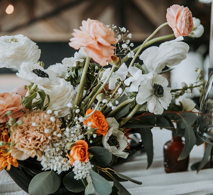 Wedding flower centrepiece decor with amber ranunculus and spray roses, fluffy white delphiniums and roses. Plus, black centred anemones and green foliage 