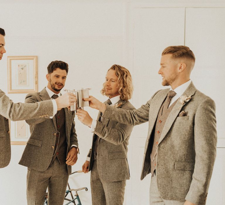 Groom and groomsmen all in sage green three piece suits raise their silver hip flasks to one another before wedding at Anran Devon