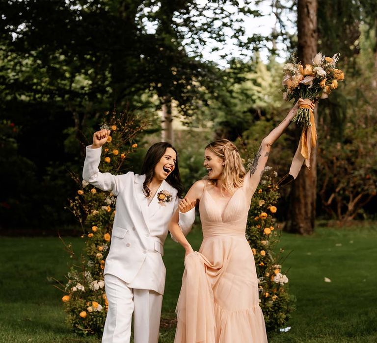 Bride in a white suit and blush pink wedding dress exiting their outdoor wedding ceremony as wife and wife 