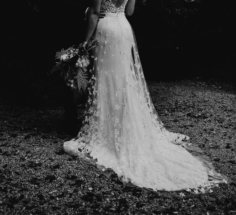 Bride in open back Rime Arodaky wedding dress with lace train holding dried bridal bouquet stand sin garden with groom in blue suit after summer wedding ceremony in Dorset