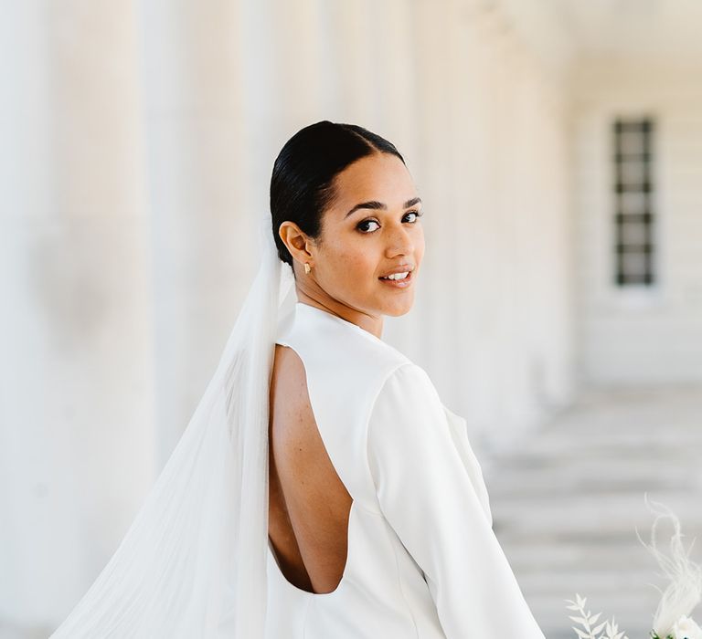 A bride wearing a backless wedding suit looks over her shoulder. She carries black and white wedding flowers.