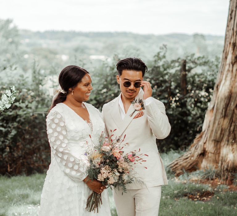 Bride in long mesh sleeve spotted Vagabond wedding dress and bridal hair bow holding mixed bridal bouquet looks at groom in white shawl collared shirt and double breasted linen suit wearing round sunglasses as they stand in the grounds at Wasing Park wedding