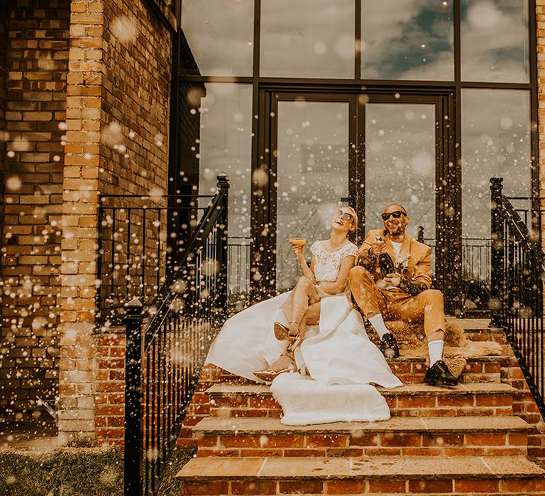 Bride in a Jesus Peiro wedding dress and groom in a mustard suit popping champagne on the steps at Curdshall Barn in Norfolk 