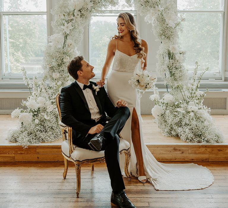 Portrait of the groom in a black tuxedo sitting on a chair with his bride in a fitted wedding dress with front split and embellished bodice in front of a white floral arch