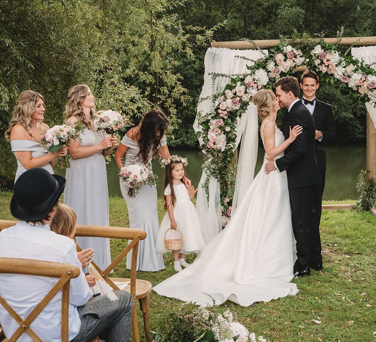 Groom in a black tuxedo kissing his bride in a Stella York wedding dress during the outdoor wedding ceremony with white and pink floral arch 