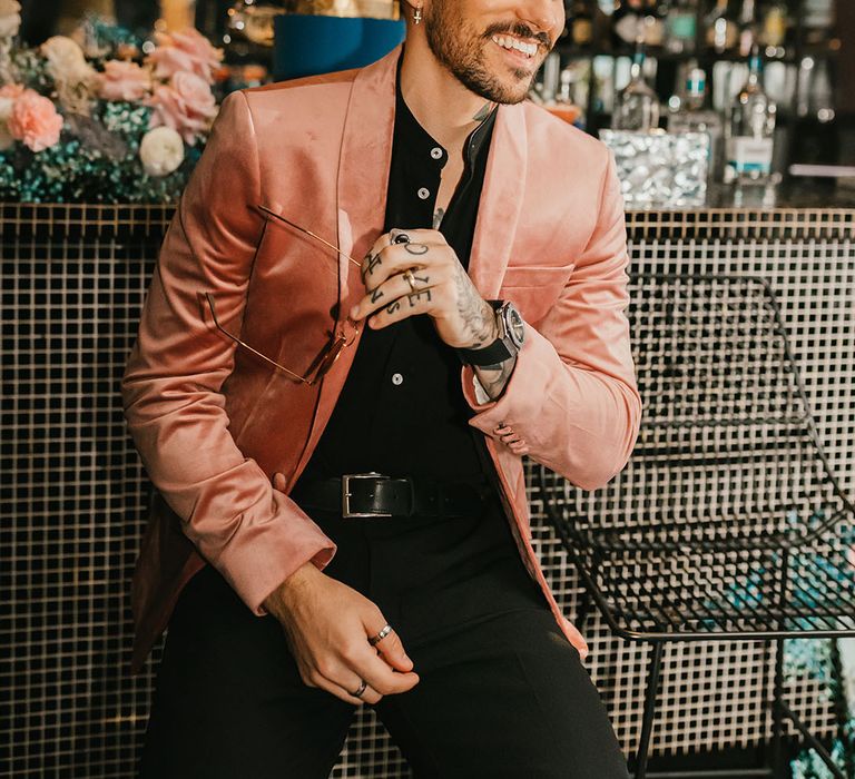 Stylish groom in a black shirt and pink velvet jacket sitting at the bar 