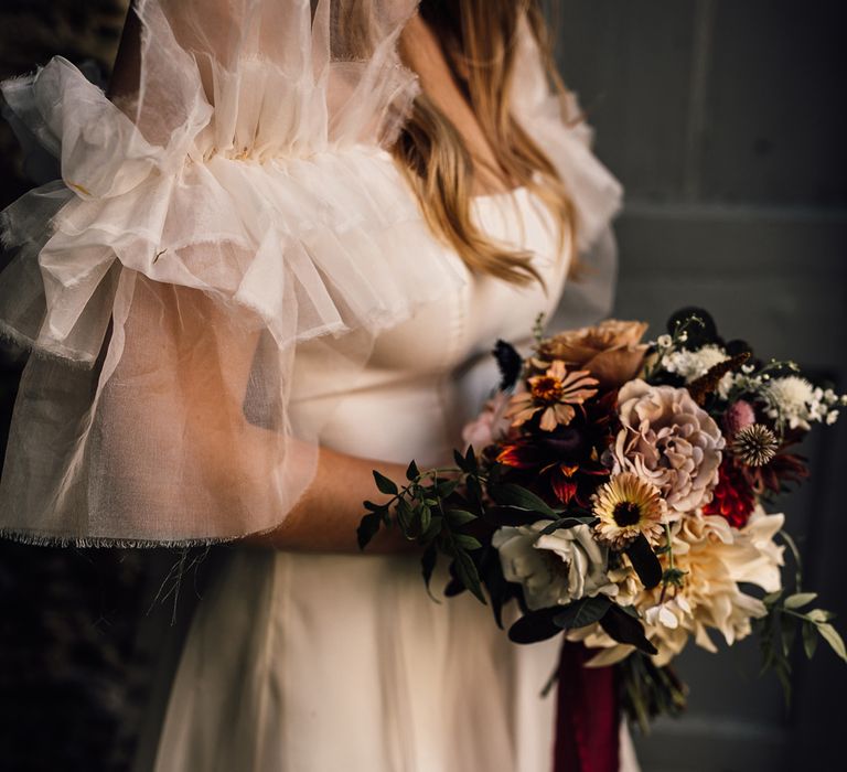 Bride in white Halfpenny London Mayfair dress with ruffle sleeves holding mixed bridal bouquet with burgundy ribbon at Cornwall wedding