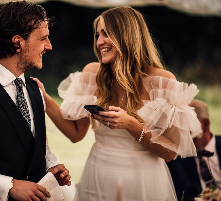 Groom in white shirt, blue waistcoat and blue patterned ties smiles at bride in white Halfpenny Mayfair London dress as she makes speech during garden marquee wedding reception in Cornwall 